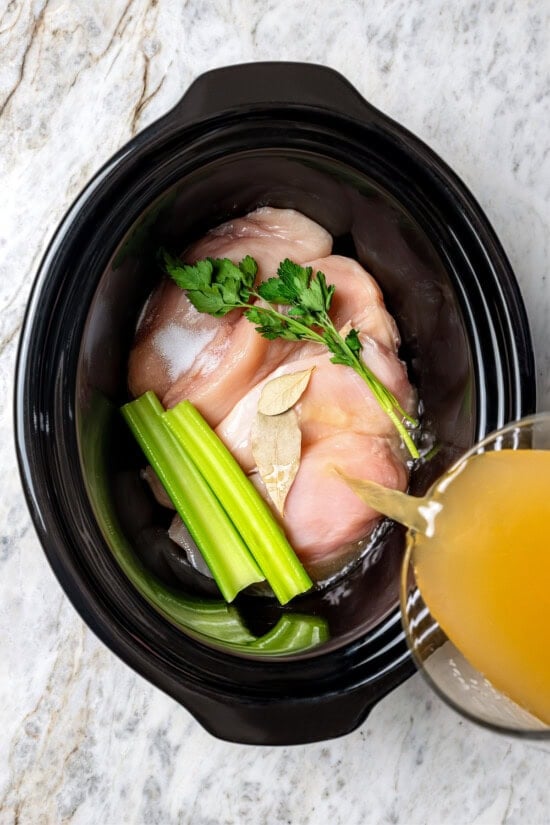 Pouring broth into slow cooker with chicken breasts, parsley, celery, and bay leaves