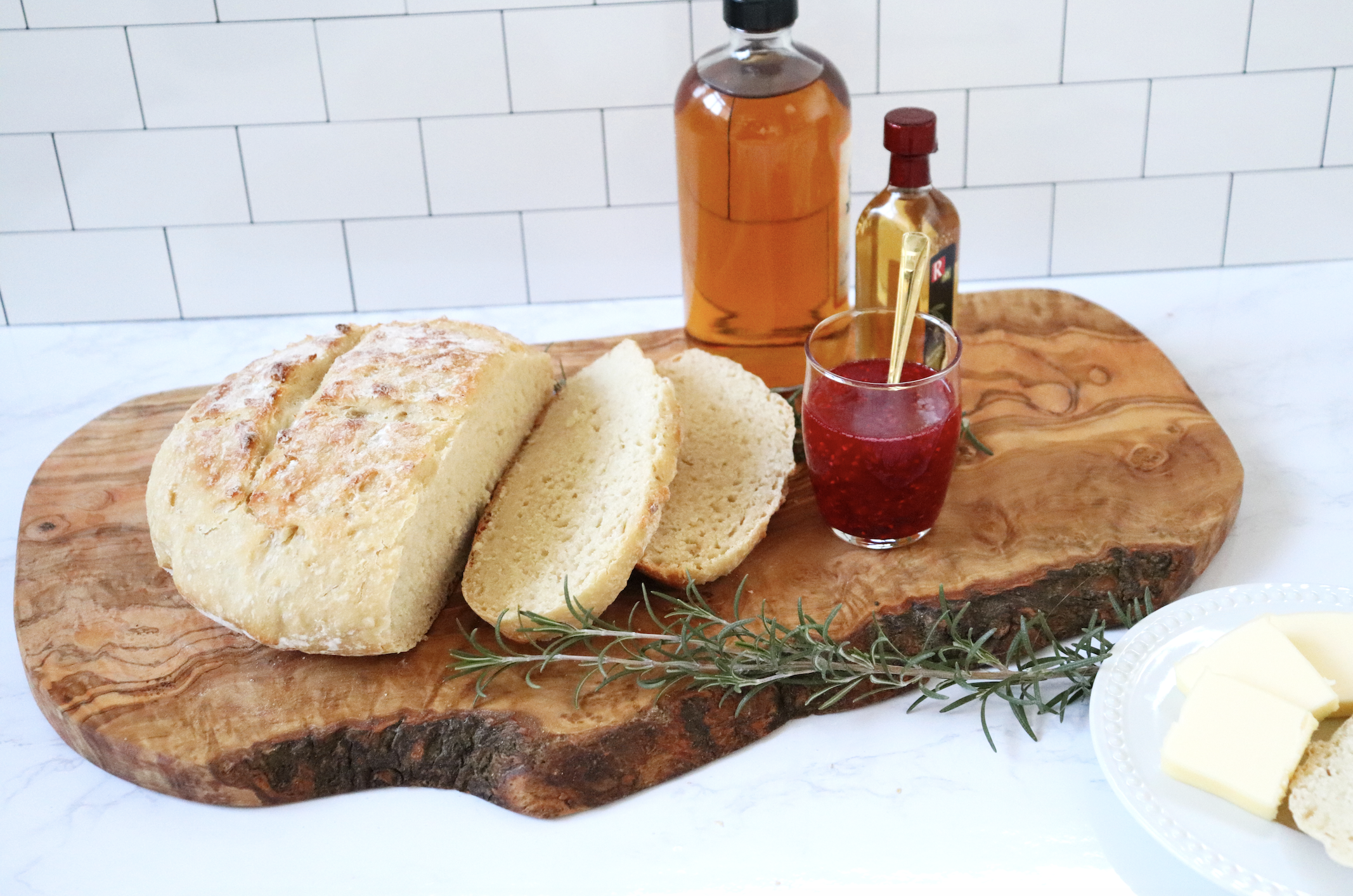 Sliced dutch oven bread, a jar of jam, and dipping oil on a wooden board with a sprig of rosemary.