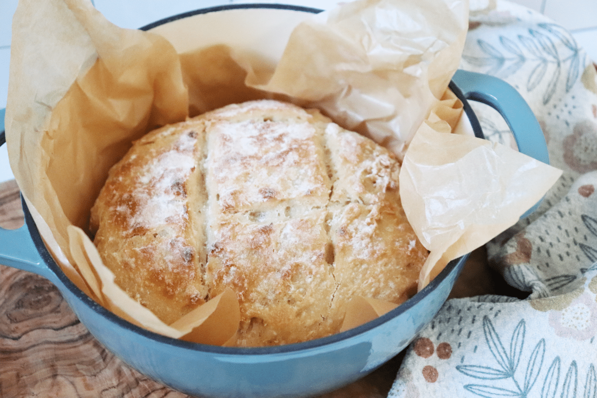 A loaf of homemade artisan bread in a blue pot with a ruffle of parchment paper on a wooden cutting board.