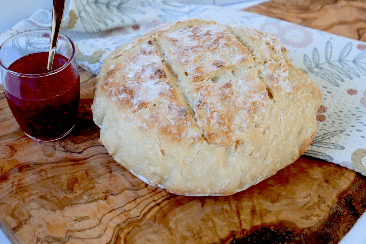 A loaf of homemade bread on a wooden board next to a jar full of jam and a flowered dish cloth