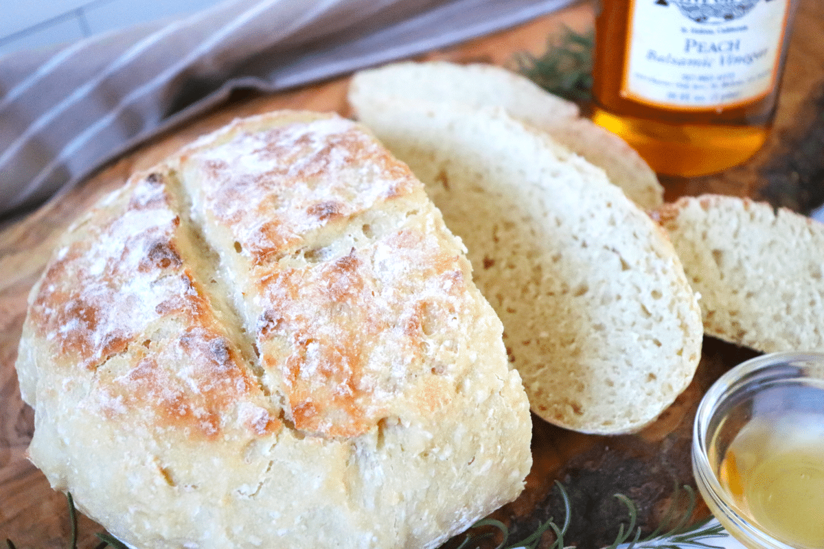 A loaf of artisan bread cut into slices on a wooden cutting board with a bowl of dipping oil.