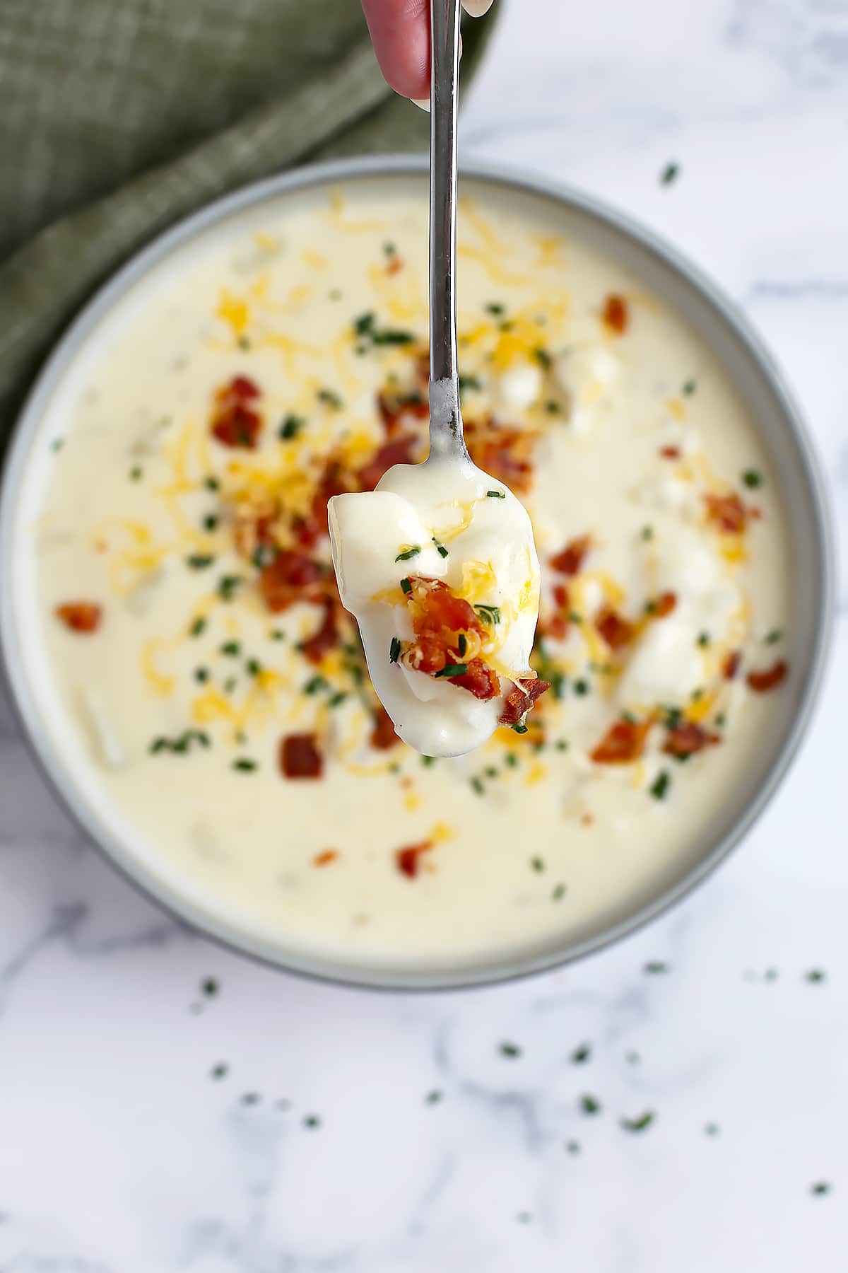 A spoon loaded with potato soup, topped with bacon bits, chives and cheddar cheese.