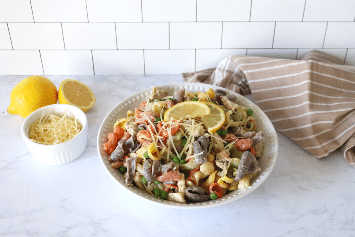 A large bowl of pasta with chicken and green peas, topped with parmesan cheese and lemon slices with a brown linen in the background.