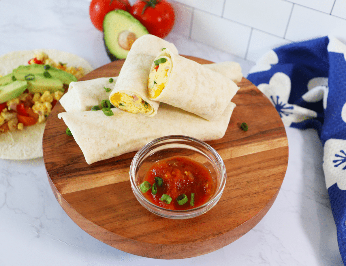 Breakfast burritos stacked on a wooden platter with a bowl of salsa topped with sliced green onions.