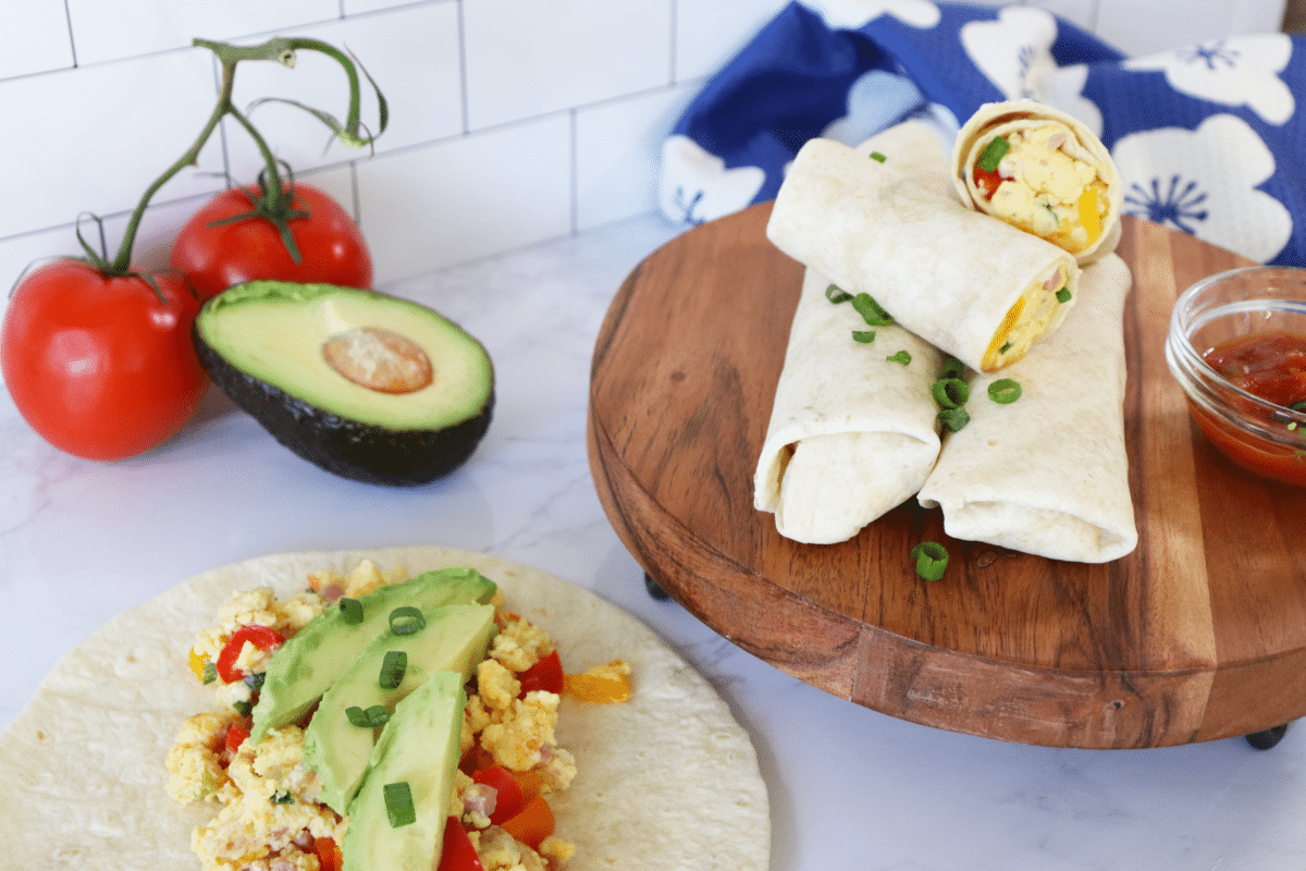 A tortilla topped with eggs, avocado and green onions with rolled burritos in the background.
