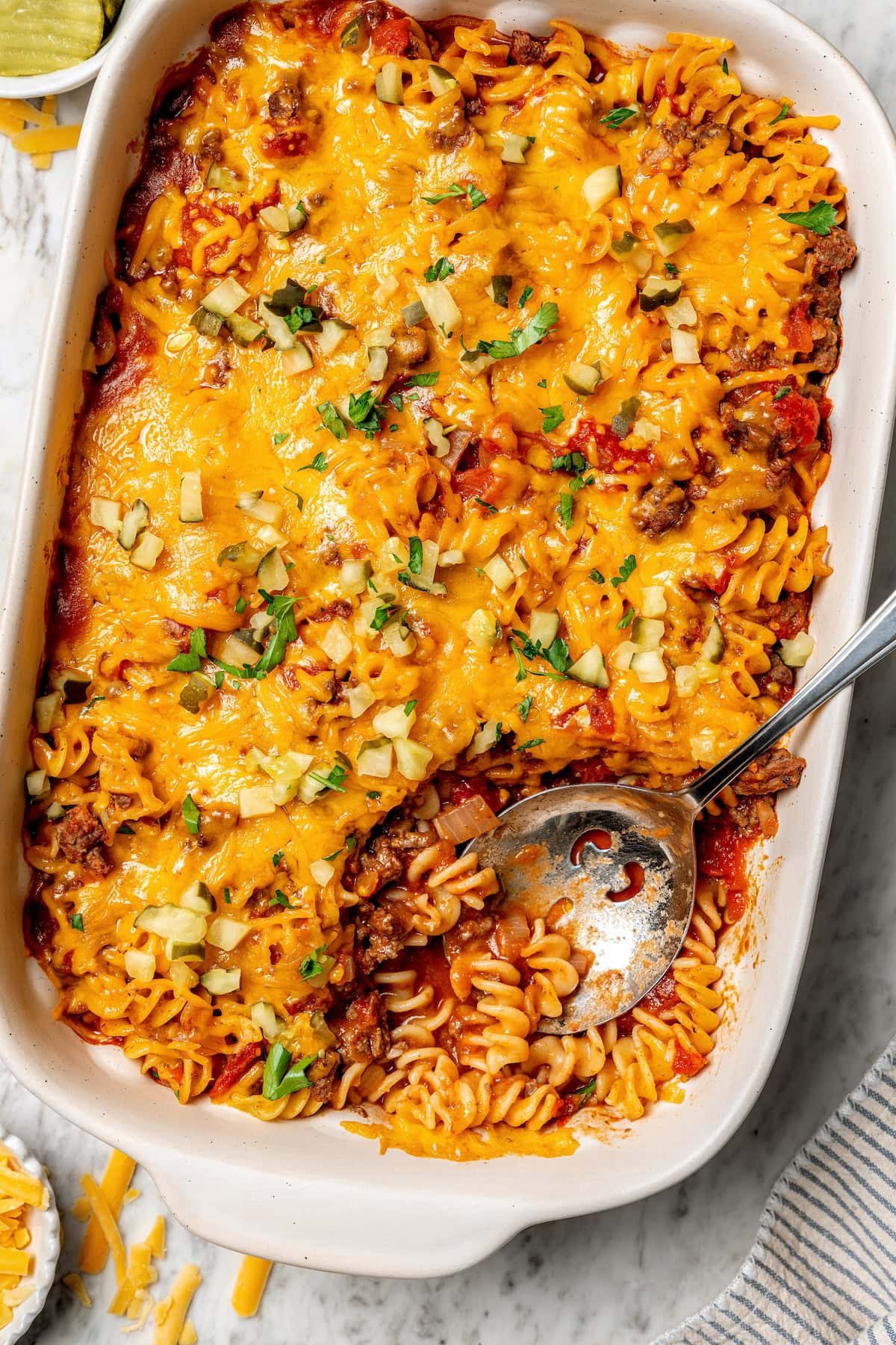 With ground beef, tomatoes, pickles, and cheese, this lightened up cheeseburger casserole recipe is kid-friendly and so delicious! Overhead view of cheeseburger casserole in baking dish with serving spoon