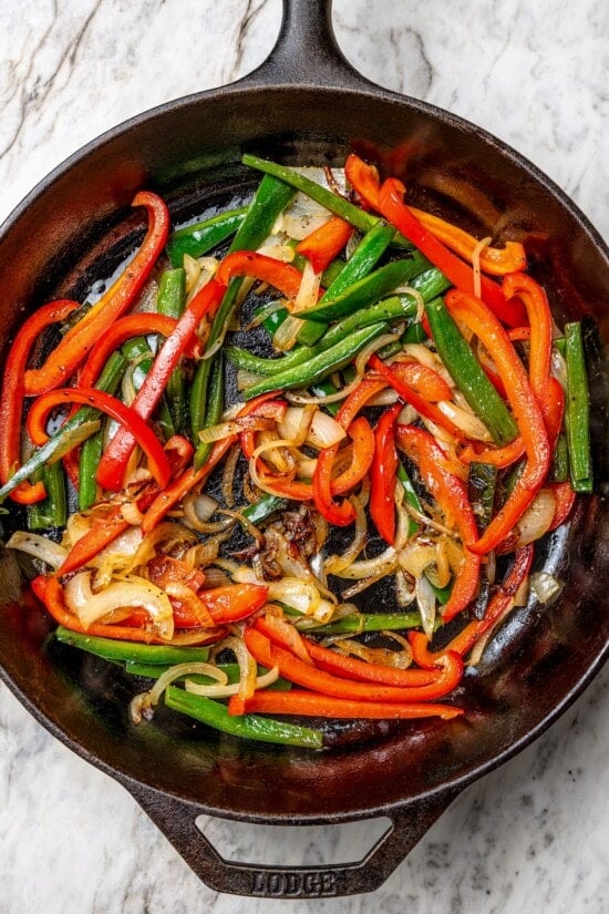 These easy chicken fajitas are made with grilled chicken breast, bell peppers and onions and served sizzling hot with tortillas and cheese! Overhead view of peppers and onions in cast iron skillet