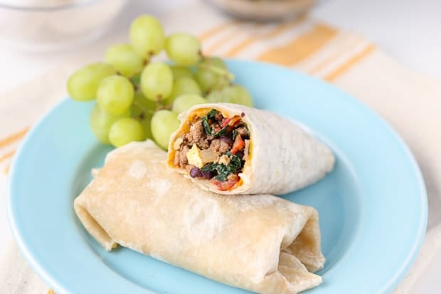 A breakfast burrito with spinach, sausage and bell peppers on a small serving plate with a side of grapes.