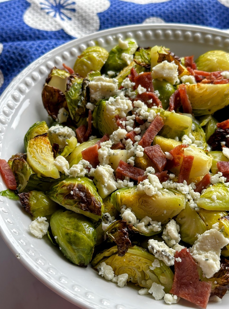 Roasted brussels sprouts on a serving platter topped with bacon crumbles and blue cheese.