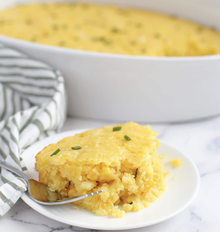 A serving of cornbread casserole with fresh corn and chives on a small serving plate with a fork and a striped linen.