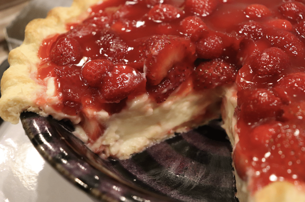 A fresh strawberry and cream pie with a slice taken out in a glass pie plate.
