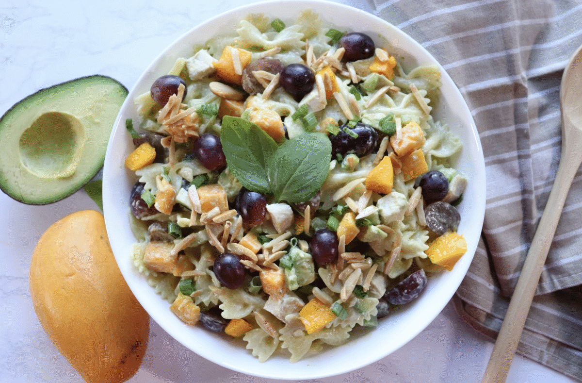 A pasta salad with grapes, mango and chicken in a serving bowl with a wooden spoon, fresh avocado and striped linen in the background.