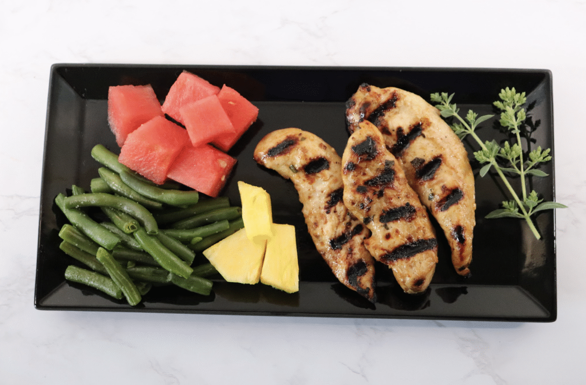 A black serving platter with grilled marinated chicken, green beans, and diced fruit.