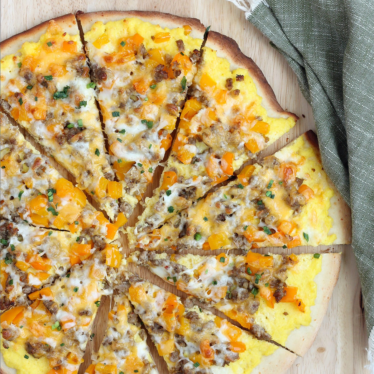 A homemade breakfast pizza topped with eggs, bell peppers, sausage and cheese on a wooden cutting board.