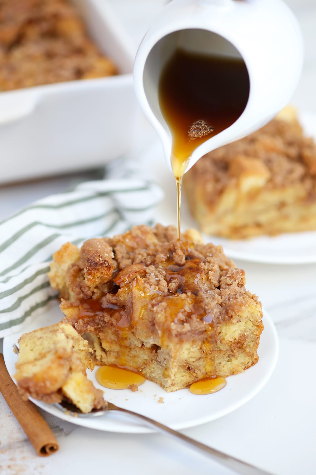 A serving of french toast casserole on a white plate drizzled with maple syrup with a striped linen in the background.