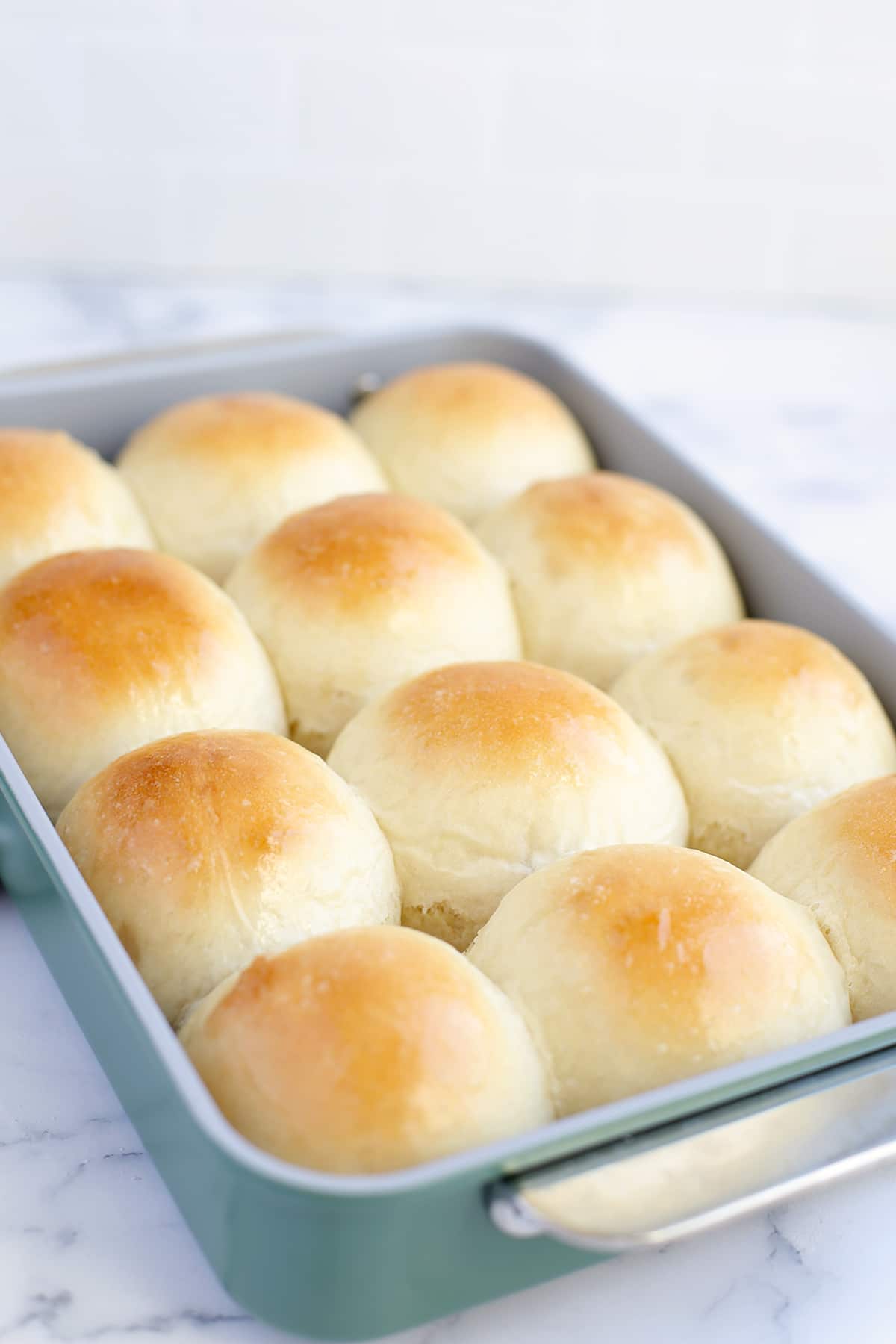 A close up shot of golden brown homemade dinner rolls in a green metal baking pan on a marble countertop. 