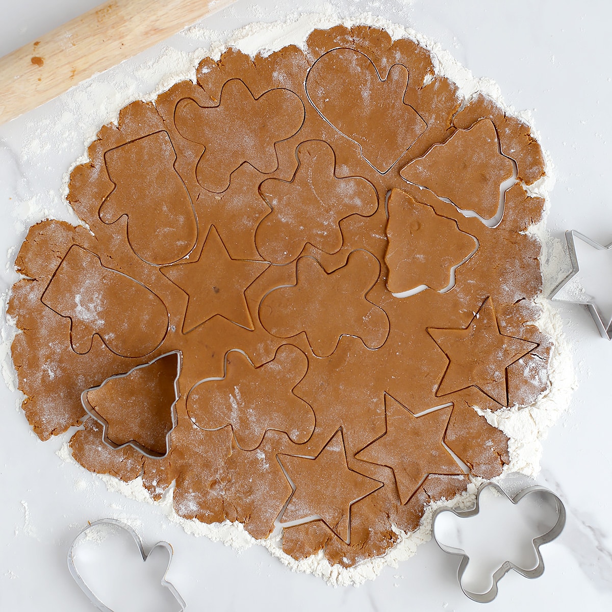 A sheet of gingerbread cookie dough rolled out on a lightly floured counter and cut into Christmas shapes.