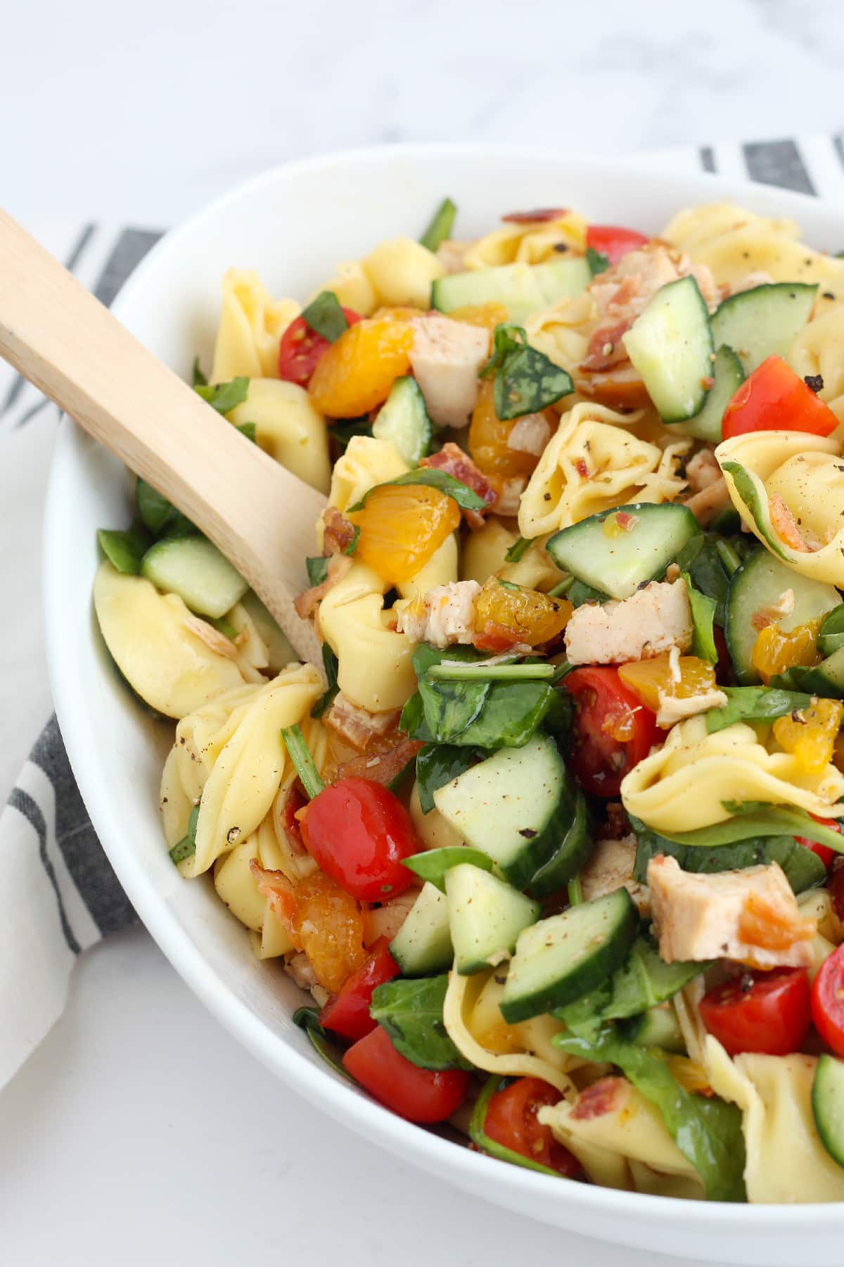 Tortellini pasta salad with cucumbers, oranges and chicken in a large bowl with a wooden serving spoon.
