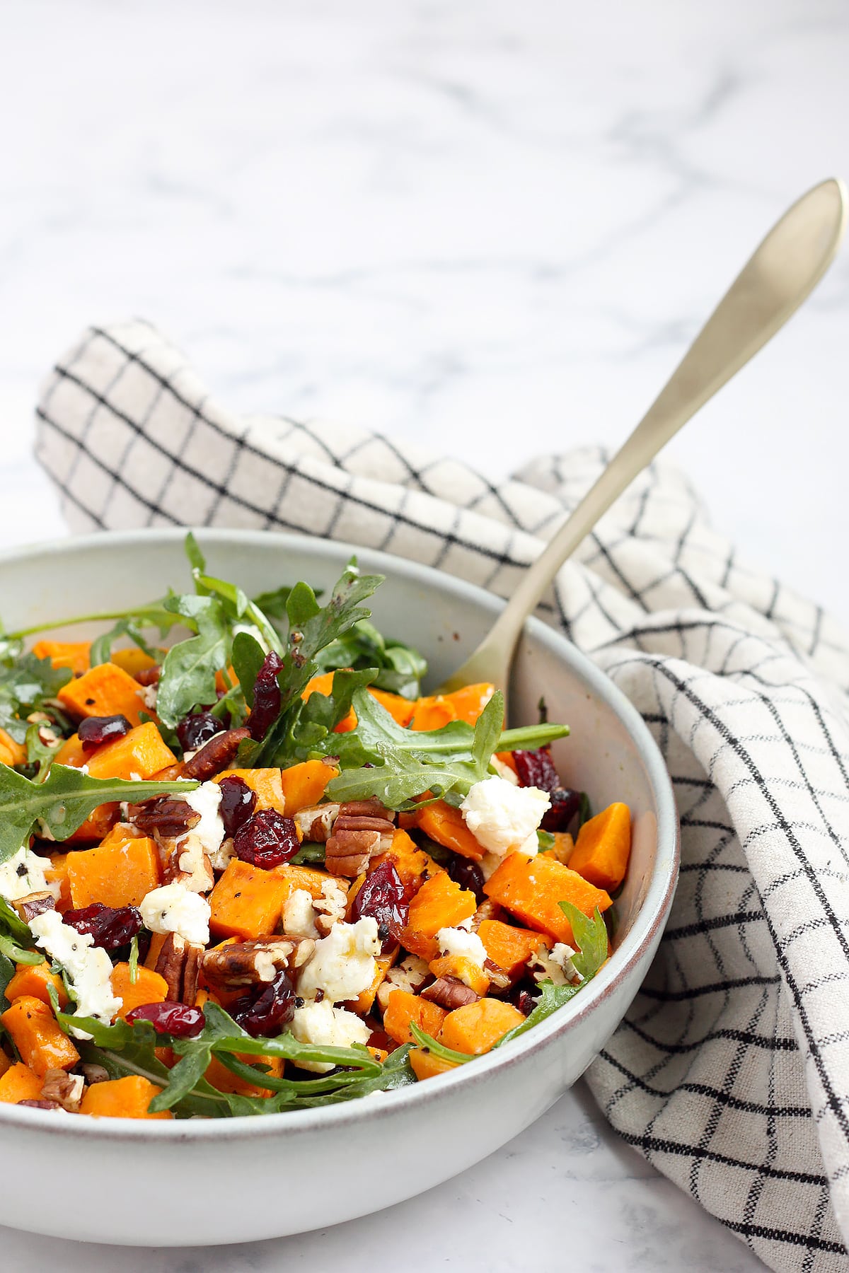 A large ceramic serving bowl with sweet potatoes, arugula, pecans, and goat cheese with a checked linen.